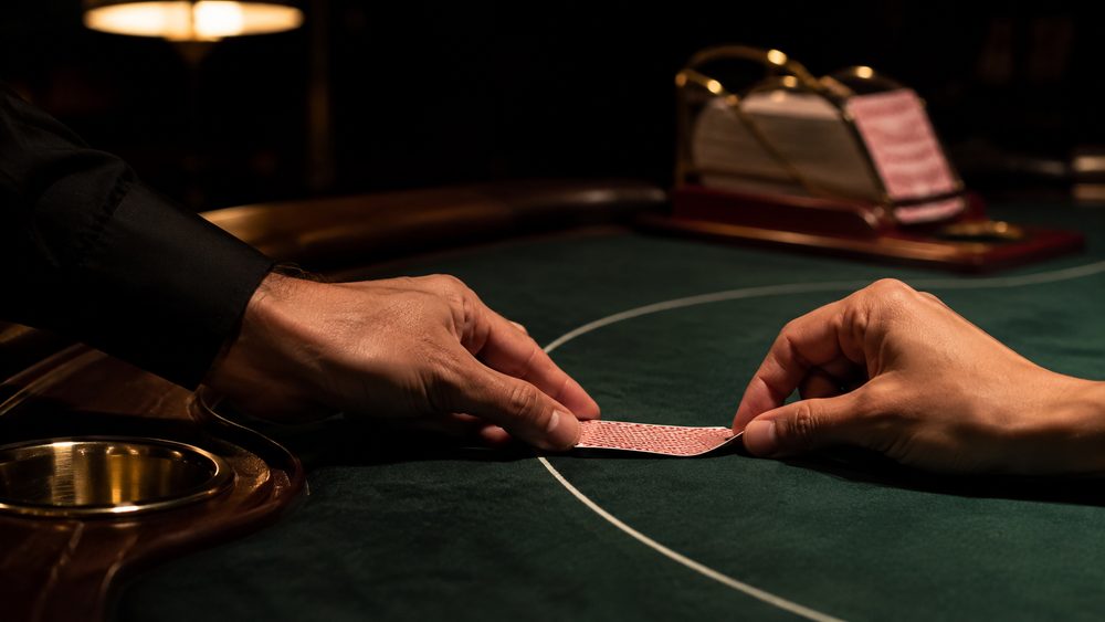 A baccarat table in a quiet high-limit room, a card being squeezed at the corner with the eight-deck shoe behind it