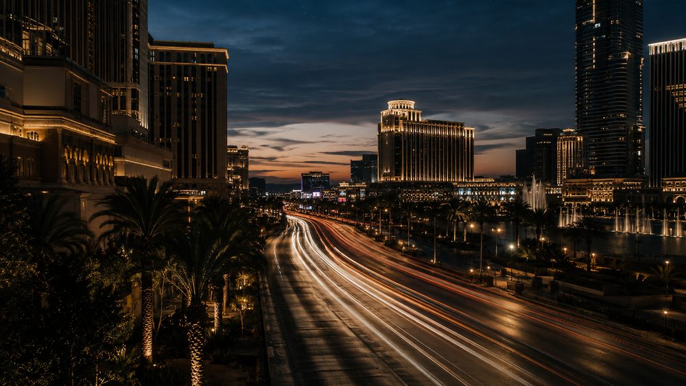 The Las Vegas Strip at blue hour from street level, neon beginning to dominate while daylight remains in the sky, traffic rendered as light trails