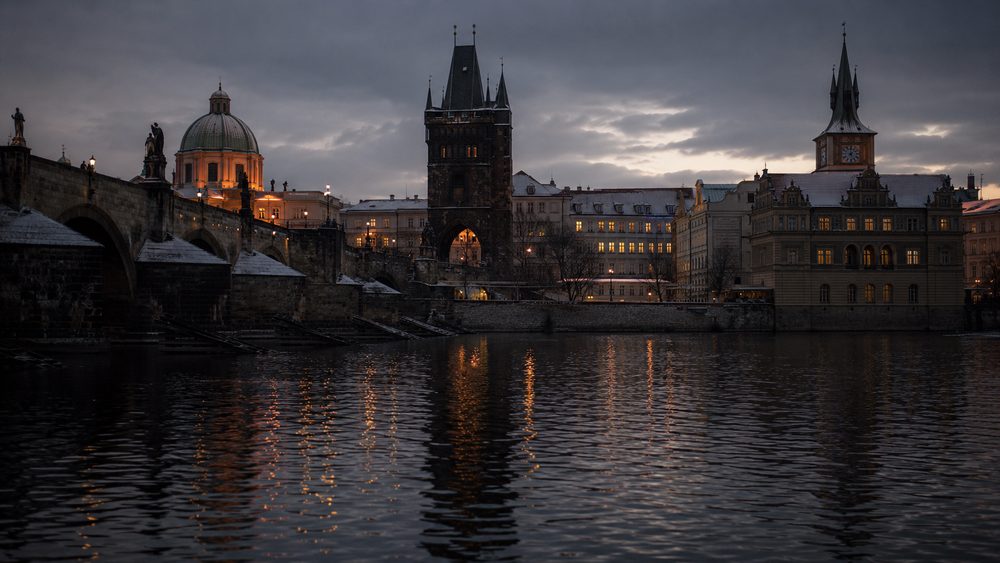 The Vltava river at dusk with Charles Bridge and the Old Town skyline, window lights beginning to come on